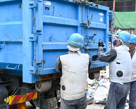 青い荷台のトラックの後ろに、3人の作業員が集まって荷台の留め具を操作しているところ